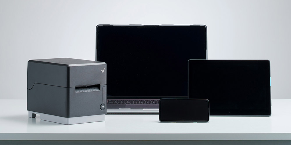 iOS端末・Android端末・PCのいずれからも柔軟に運用できるラベルプリンター mC-Label3シリーズ（MCL32CI / MCL32CBI）とタブレット・スマートフォンなどのスマートデバイスと並んで置いてある画像。